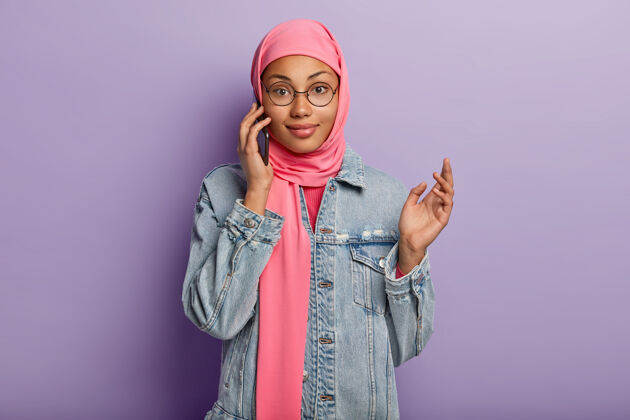 女性上身镜头享受智能手机对话 是现代设备的高级用户 戴粉色头巾和牛仔夹克 使用公共互联网连接 隔离在紫色墙上伊斯兰智能手机小玩意