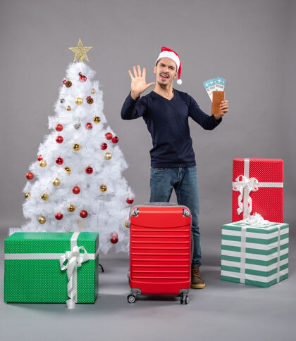 五满意的人拿着红色的手提箱 拿着他的旅行票 向灰色的击掌红色圣诞树高