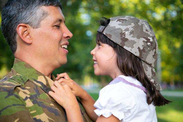 父亲快乐正面的父亲抱着小女儿 抱着小女孩 在军事任务结束后和她在户外交谈特写镜头家庭团聚或回家的概念黑色伪装女儿