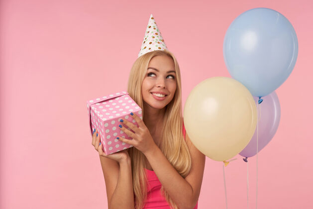情感身着粉色上衣 头戴生日帽的快乐金发女郎的肖像 在五彩气球中摆姿势 开心地看着一边 手里拿着礼物盒 站在粉色背景上微笑肖像欢快