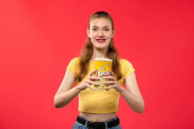 美女正面图年轻女性在电影院拿着爆米花包 微笑着看红墙电影院的小吃女性趣味电影女性爆米花电影