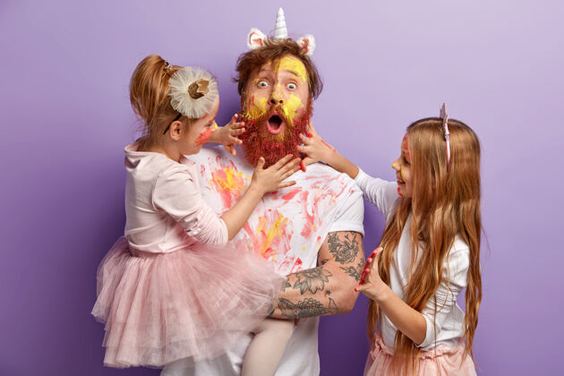 高兴惊讶的爸爸和两个女孩在家里一起玩耍 用水彩画脸 玩得开心 展示用鲜艳的颜色画的手 隔离在紫色的墙上全家福做父亲女儿水彩小