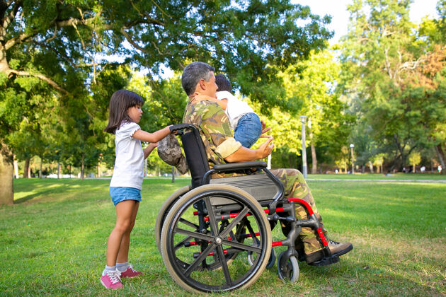 视图中年残疾军人爸爸带着两个孩子在公园散步女孩拿着轮椅把手 男孩站在爸爸腿上退伍军人或残疾概念父亲部队享受