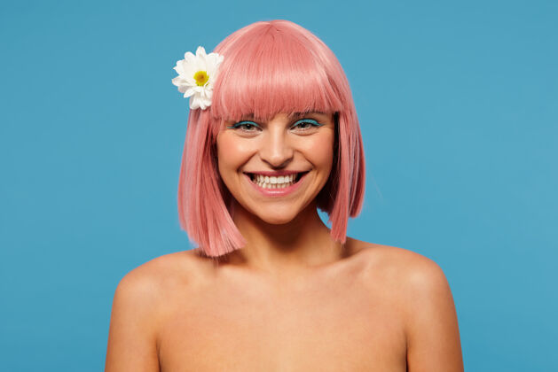 湛蓝快乐的年轻漂亮的粉色头发的女人 留着鲍勃式的发型 愉快地看着镜头 带着迷人的笑容 站在蓝色背景下 头发上插着白花优雅开朗肩膀