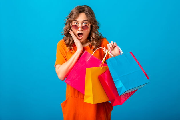 商店图片中的兴奋迷人的微笑时尚女性购物狂穿着橙色的新潮连衣裙拿着购物袋在蓝色的工作室背景隔离太阳镜女孩零售