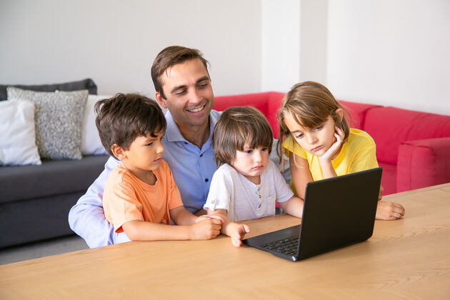 看快乐的爸爸和沉思的孩子们在周末一起通过笔记本电脑看电影快乐的爸爸和孩子们坐在客厅的桌子旁父亲 童年和数字技术的概念儿子枕头童年