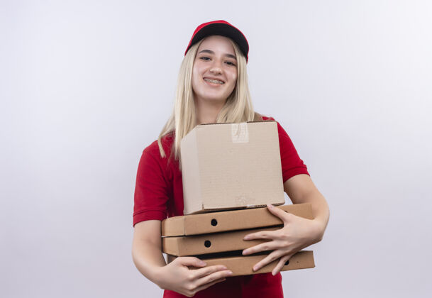 白色微笑的小女孩穿着红色t恤 戴着帽子 戴着牙套 拿着盒子和披萨盒 背景是白色的红色年轻女孩