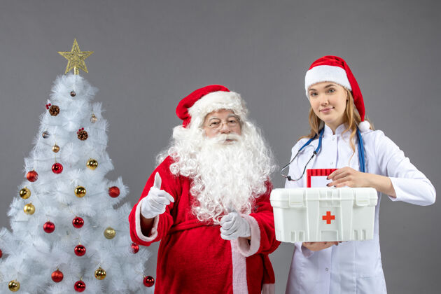 帽子圣诞老人和女医生的前视图 女医生拿着急救箱在灰色的墙上前面援助人