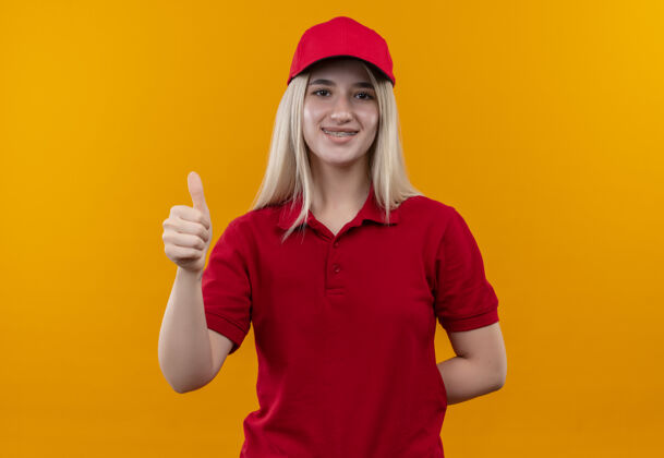 支撑微笑的小女孩穿着红色t恤 戴着帽子 戴着牙套 在孤立的橙色背景上竖起大拇指拇指帽子牙科