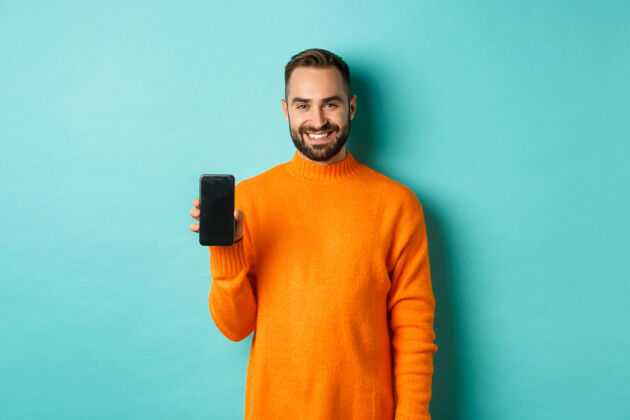 摆姿势帅哥留着胡子 穿着橘色毛衣 露出智能手机屏幕 面带微笑 在网上展示宣传片 绿松石墙模型现代使用