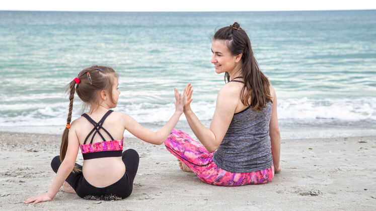快乐的家庭一位年轻的母亲带着一个穿着运动服的小女儿坐在海边家庭价值观和健康的生活方式夏天女儿家庭