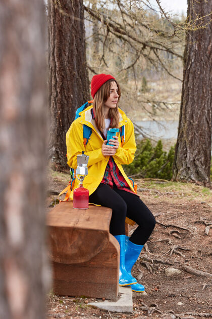 自然图为体贴的女游客在森林里的木凳上休息 用热水瓶喝茶 在野营的炉子上煮咖啡 戴着红帽子饮料茶紧身裤
