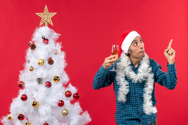 玻璃圣诞心情快乐的年轻人戴着圣诞老人的帽子 穿着一件蓝色的衬衫 举起一杯葡萄酒 在圣诞树附近展示上面的画面新年前夜年圣诞老人