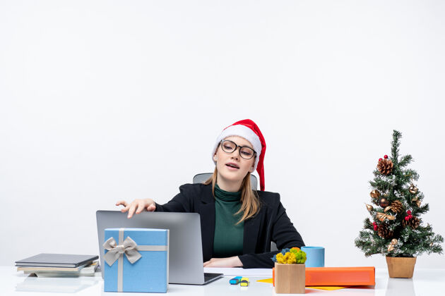 圣诞老人一位年轻漂亮的女士戴着圣诞老人的帽子坐在桌旁 桌上放着圣诞树和一份礼物 她在办公室里合上了她的笔记本笔记本电脑帽子圣诞节