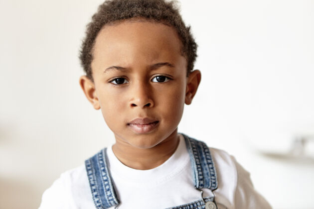 调皮脾气暴躁可爱的非洲小男孩穿着牛仔连身衣和白色t恤室内孩子肖像