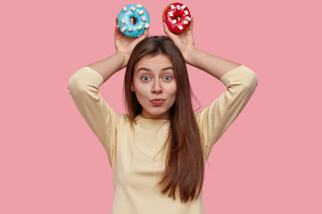 食物可爱有趣的黑发年轻女子拿着两个美味的甜甜圈 穿着黄色的衣服 模特在粉红色的空间个人面包店零食