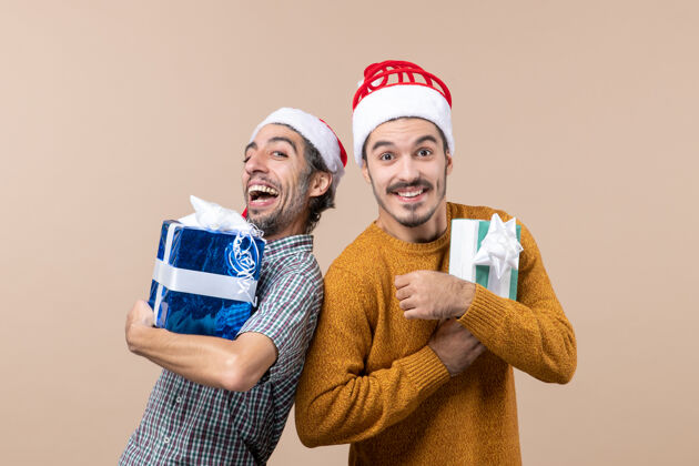 男性正面图两个朋友背靠背摆姿势拿着圣诞礼物在米色孤立的背景两个朋友成人