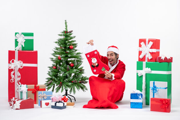 圣诞老人新年气氛有趣积极的圣诞老人坐在地上 展示圣诞袜子附近的礼物和装饰圣诞树上的白色背景圣诞老人圣诞树袜子