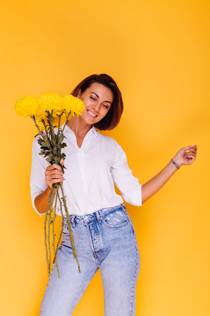 华丽摄影棚拍摄的黄色背景快乐的白人妇女短发穿着休闲服白衬衫和牛仔裤手持一束黄色紫苑微笑年轻人