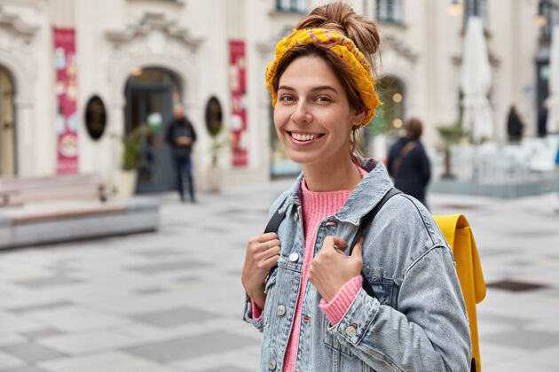 满意积极乐观的女人有快乐的表情 穿着时髦的衣服 背着黄色的小背包 游荡在城市的街道上学生头带行人