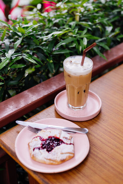 棕色木制桌子上粉红色的架子上放着冰拿铁 夏天咖啡馆绿树丛中放着小红莓馅饼全麦奶油谷物