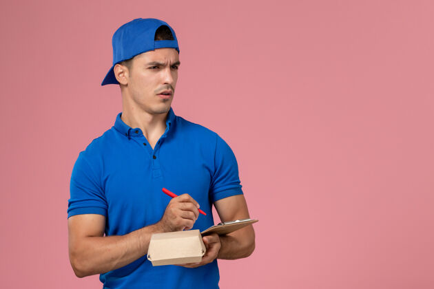 视图正面图身穿蓝色制服披肩的年轻男性快递员拿着小快递食品包和记事本在浅粉色的墙上写笔记食物制服递送