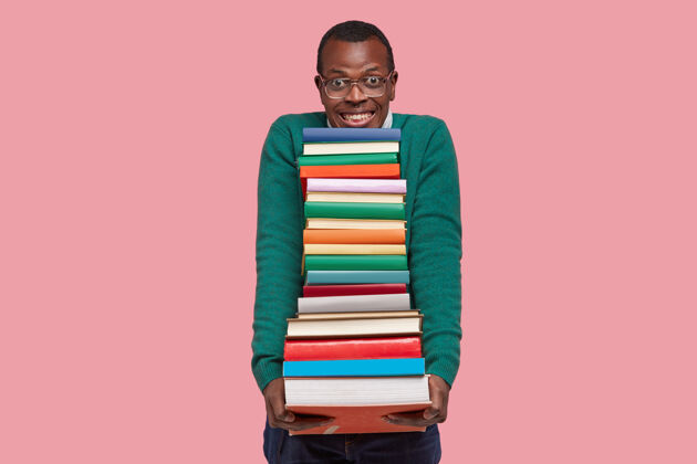 积极积极向上的非裔美国人手里拿着一大堆教科书 笑容满面 戴着眼镜和绿色毛衣 隔离在粉色背景下 准备上课男孩高兴堆