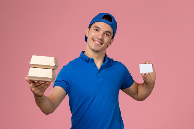 送货正面图：身穿蓝色制服披肩的年轻男性快递员拿着小包裹 浅粉色墙上有卡片微笑服务食物