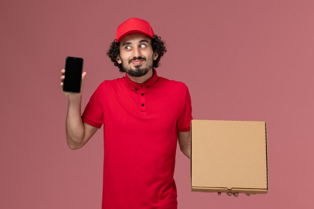 肖像正面图穿着红色衬衫和披风的男快递员拿着空的快递食品箱 粉色墙上挂着电话电话空工人