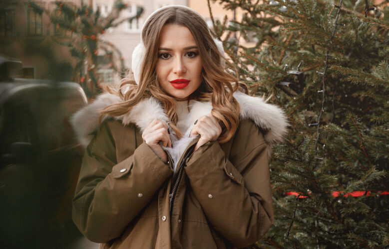 圣诞树街上圣诞树旁穿着皮草夹克的时髦女孩戴着温暖的耳机和飘逸的头发的年轻女子耳罩或冬季耳机冬天雪皮草大衣人