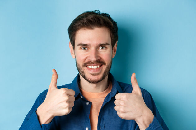 表情笑容可掬的帅哥竖起大拇指 称赞工作 推荐公司 站在蓝色背景上的特写镜头休闲时尚年轻