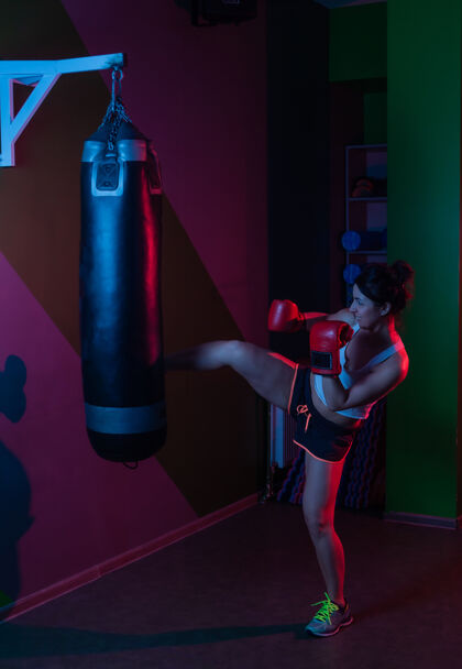 拳击手戴拳击手套的女拳击手在黑暗背景下用红蓝色霓虹灯踢拳击袋活跃力量拳击