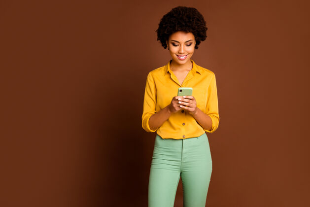 年轻照片惊人的深色皮肤波浪女士握着电话的手影响力写新的博客帖子创意青年主题穿黄色衬衫绿色裤子孤立棕色美国人影响者情感