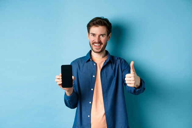 休闲帅气的年轻人竖起大拇指 空着智能手机屏幕 推荐应用程序或公司 站在蓝色背景上发型年轻社交