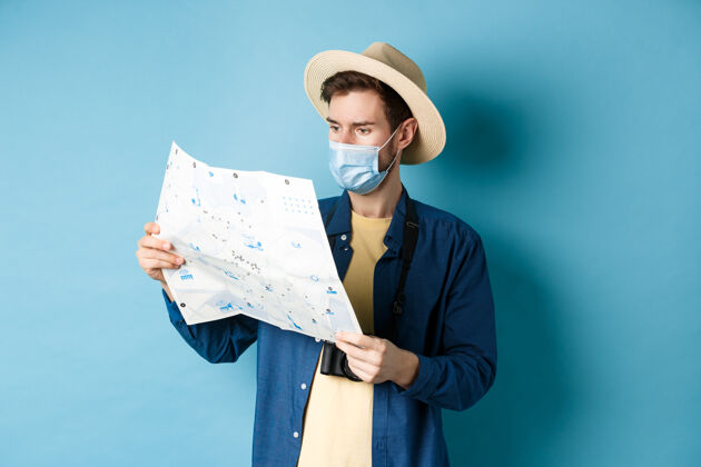 酷Covid-19 流行病和旅行概念戴着防暑帽 戴着口罩的帅哥 看着旅游地图 旅游度假 站在蓝色的背景上冠状病毒医学成人