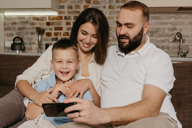 电话一位留着胡须的幸福丈夫正在智能手机上向一对欢笑的孩子和一位妻子演示这个节目沙发肖像男性