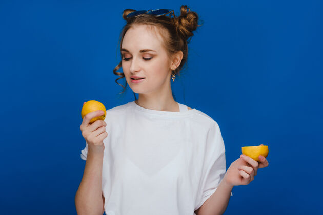 柠檬一个年轻漂亮的女孩站在蓝色的背景上 手里拿着柠檬 咬着嘴巴饮食食物