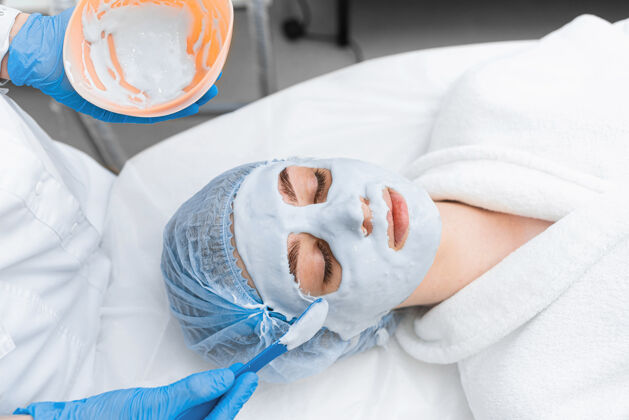技术一个穿着白色长袍的女孩脸上戴着海藻酸盐面具面部美容院治疗