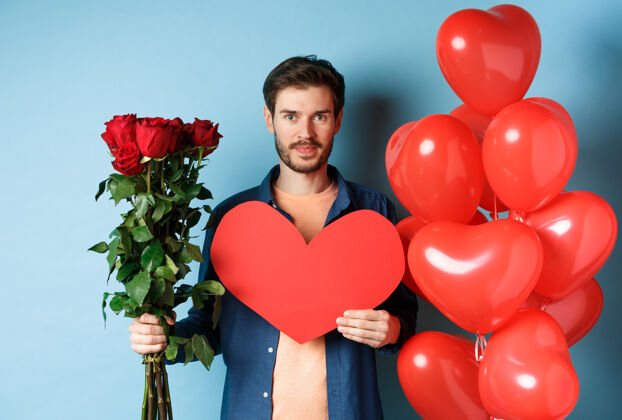 微笑情人节浪漫带着一束红玫瑰和心形气球的年轻人微笑着 站在蓝色背景上 为情人节的约会带来礼物男朋友男人丈夫