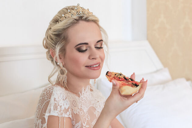 婚礼日微笑的金发新娘在酒店房间里吃比萨饼衣服酒店脸