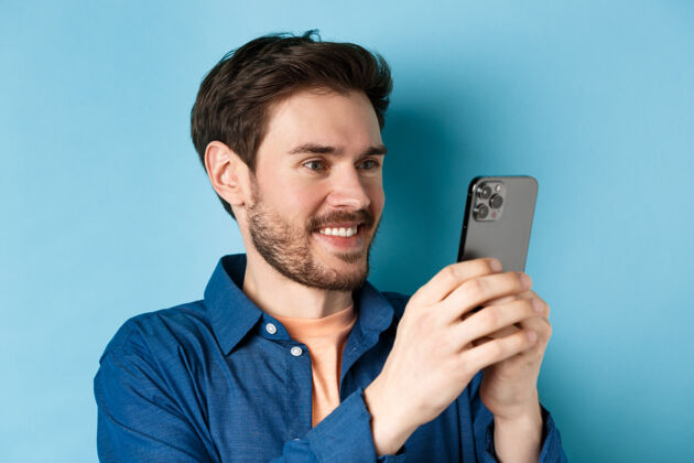 社交一个英俊的白种人站在蓝色的背景上 一边阅读手机屏幕 一边用智能手机拍照 一边微笑表情网络手机
