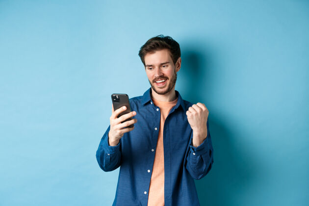 网络幸运的家伙在网上赢了 看手机屏幕和拳头泵 胜利 站在蓝色的背景上的形象肖像手机休闲