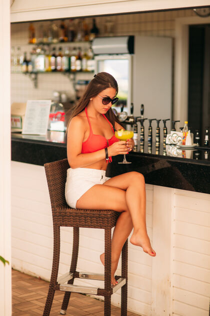年轻美女在沙滩酒吧休息 喝一杯清爽的鸡尾酒鲜艳的泳衣和太阳镜夏天酒精白色