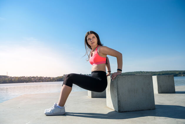 蹲在湖边阳光明媚的公园里做蹲式运动的女运动员健康的概念和健康的生活方式活跃健身女孩
