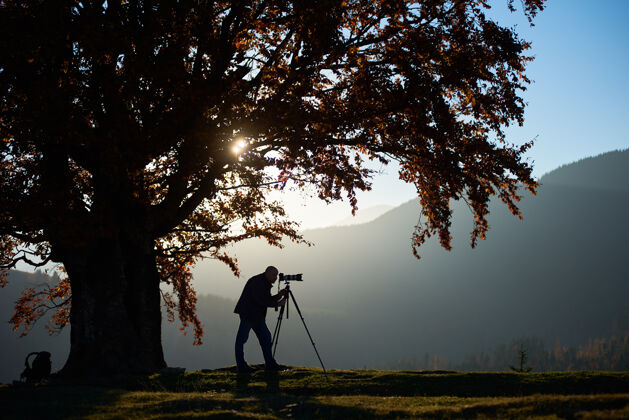 森林徒步旅行者带着相机在大树下的山景背景下的草谷里风景设备成人