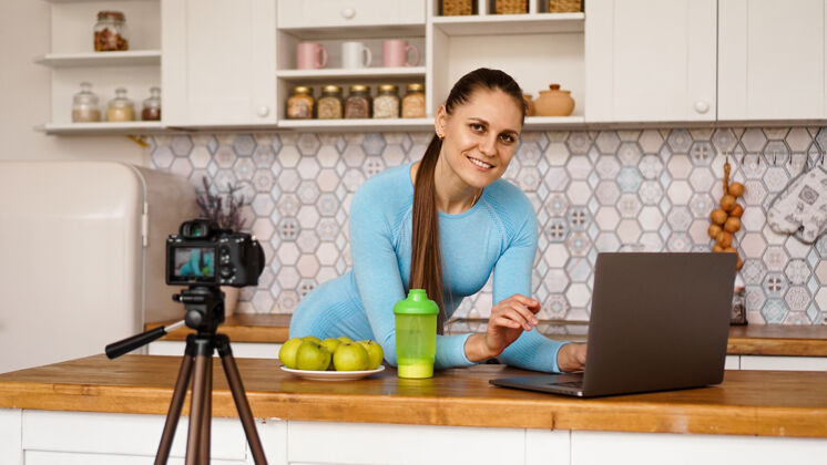 网站厨房里拿着笔记本电脑的年轻女子微笑着美食博客的概念一位女士正在录制一段关于健康饮食的视频三脚架上的相机电脑记录博客
