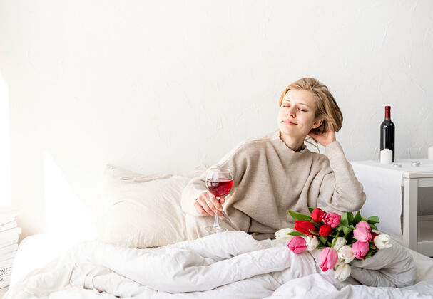生活方式快乐的女人穿着睡衣坐在床上 愉快地欣赏着鲜花和一杯红酒白天春天爱情