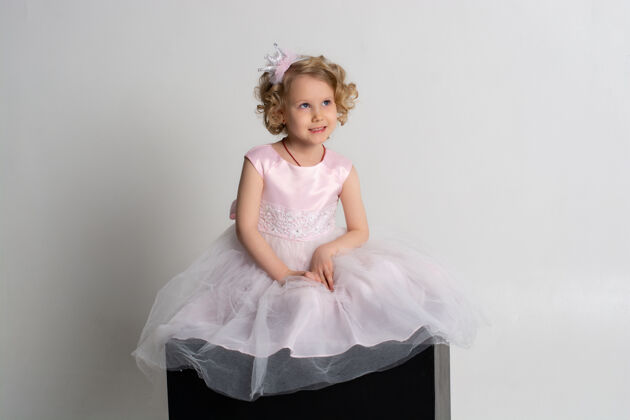 头发一个小女孩穿着粉红色的裙子 头戴皇冠 坐在白色背景的黑色立方体上年轻小美丽