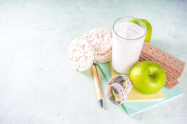 每日健康食品和减肥概念苹果 酸奶 健康的谷类食品 面包 笔记本和卷尺放在浅绿色的墙上 可以复印文本计划时间表磁带