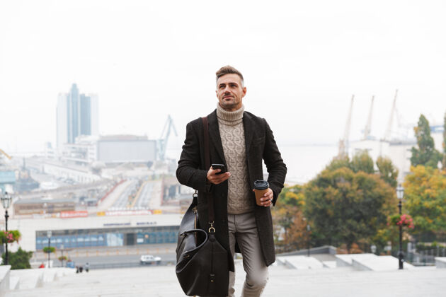 帅气30多岁的时尚男人的肖像 穿着夹克 拿着手机和外卖咖啡 走在城市街道上站立散步杯子